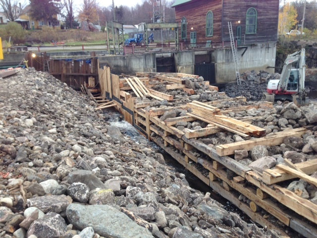 With the temporary coffer dam (on the left) diverting the St. Regis River, a local crew laid in a crib of tamarack logs stuffed and weighted with rock and boulders. (Source: NCPR)